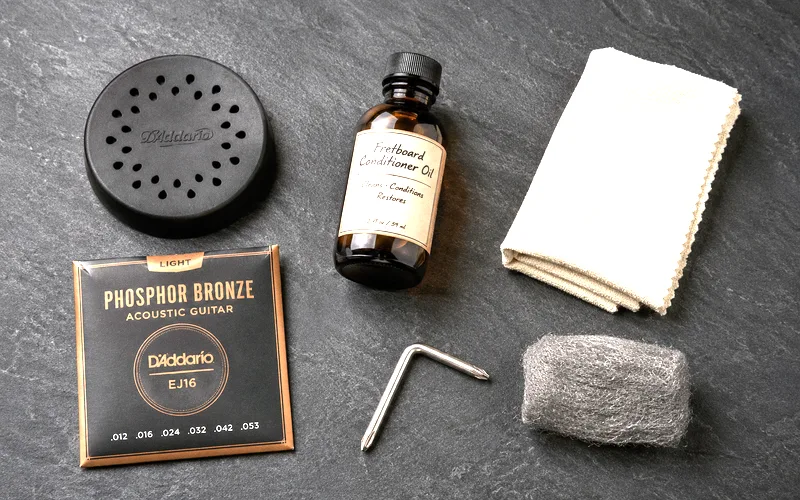 Acoustic guitar maintenance supplies including a humidifier, fretboard oil, cleaning cloth, and fresh set of strings