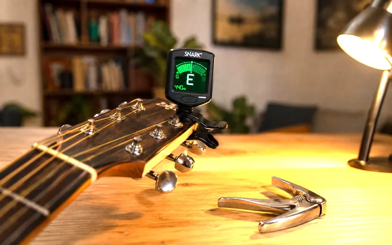 Clip-on guitar tuner attached to an acoustic guitar headstock next to a spring-loaded capo on a wooden table
