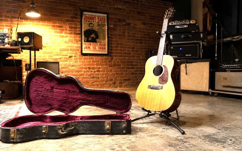 Hard shell guitar case open on the floor next to a tripod guitar stand holding an acoustic guitar against a studio wall
