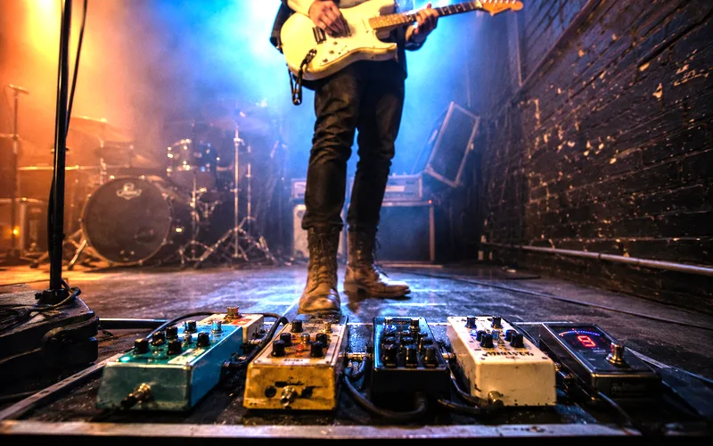 Guitarist on stage with wireless system transmitter plugged into guitar and a pedalboard with multiple effects pedals at their feet