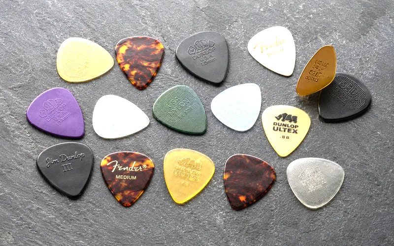 Assortment of guitar picks in different thicknesses and materials spread out on a dark surface showing thin medium and heavy gauges