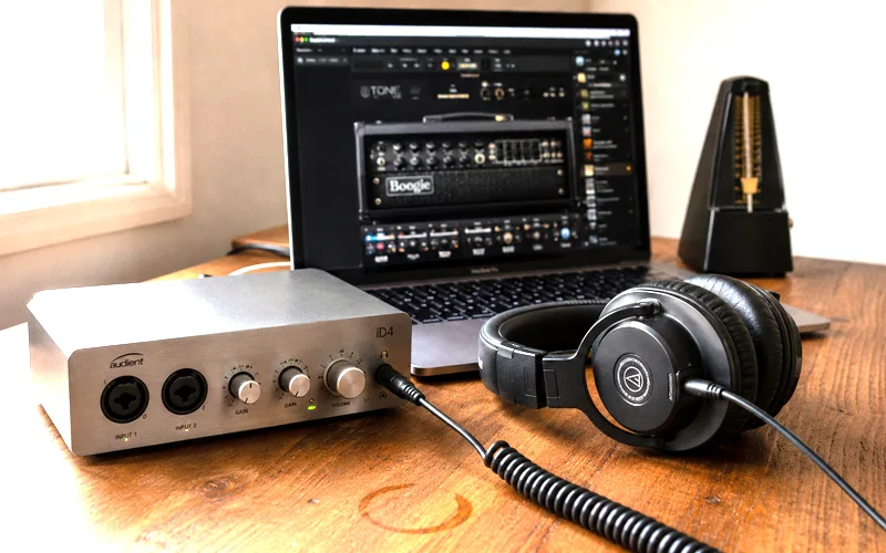Home guitar recording setup with audio interface on a desk next to closed-back headphones and a laptop running amp simulation software