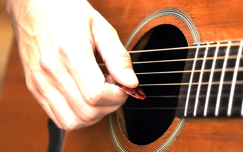 Close-up of right hand strumming an acoustic guitar with a pick near the sound hole